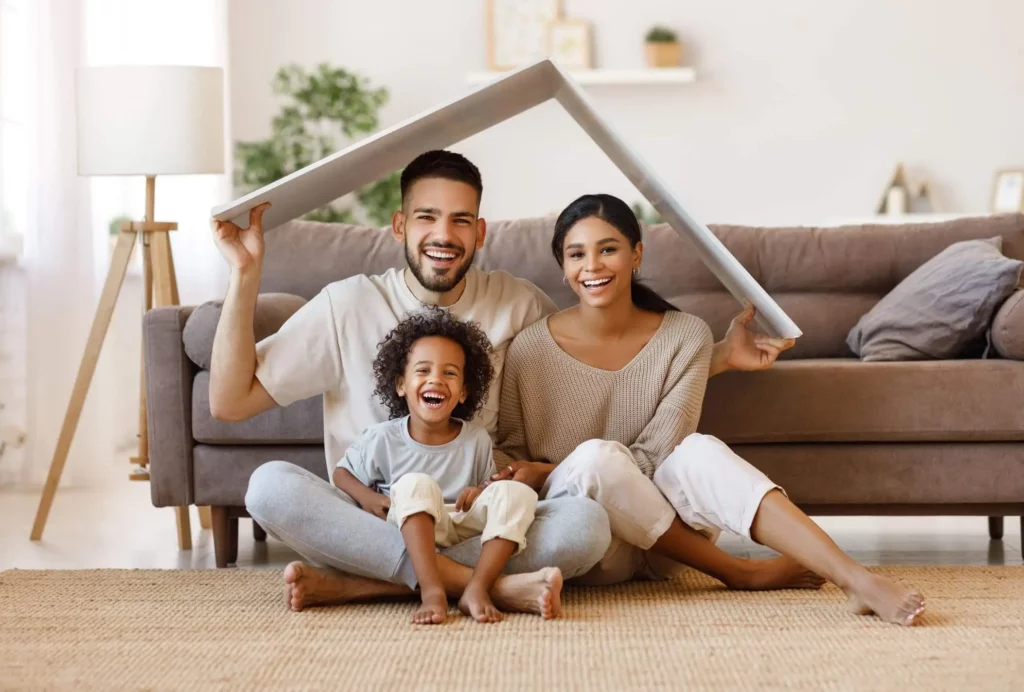 Imagem de uma família composta por um homem, uma mulher e uma criança sentados no chão da sala com um papelão em cima da cabeça simbolizando o telhado de uma casa para ilustrar matéria sobre qual o valor do condomínio
