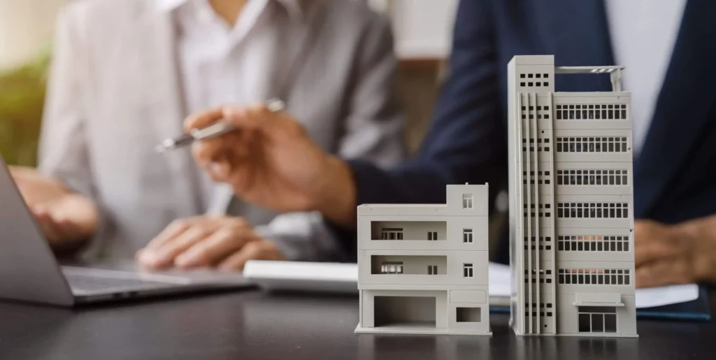 Imagem da miniatura de um prédio em cima de uma mesa, onde duas pessoas olham para a tela de um computador, para ilustrar matéria sobre valor do condomínio