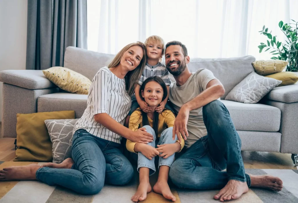 Imagem de um casal composto por um homem e uma mulher sentados sorridentes ao lado de dois filhos no tapete da sala de estar para ilustrar matéria sobre o que é IGP-M do imóvel
